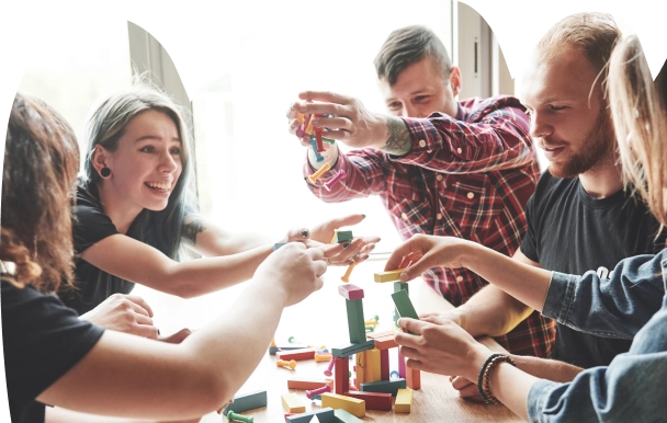 Een groep van vijf mensen bouwt vol enthousiasme een toren met kleurrijke houten blokken op een tafel, wat getuigt van teamwork en creativiteit. Helder, natuurlijk licht stroomt naar binnen.
